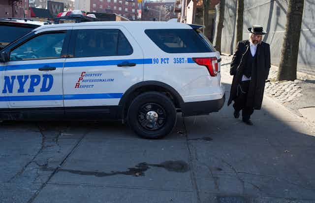 A Hasidic man with a black jacket and a hat walks behind a NYPD blue and white police car on an empty New York City street.