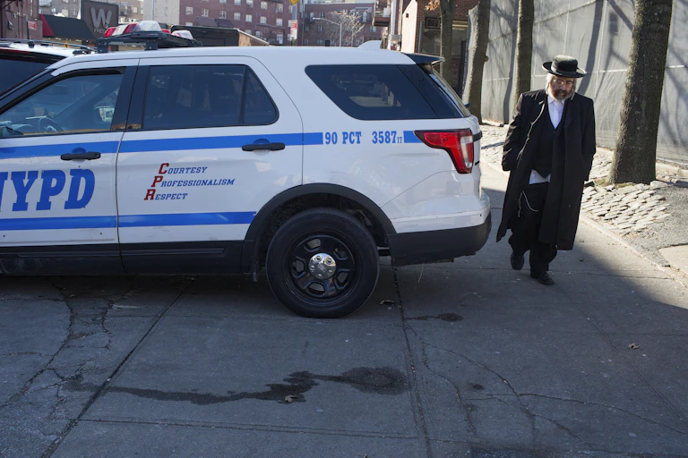 A Hasidic man with a black jacket and a hat walks behind a NYPD blue and white police car on an empty New York City street.