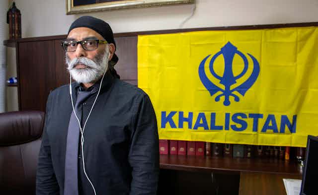 A Sikh man with a white beard stands in front of a yellow flag featuring a  khanda above the word Khalistan