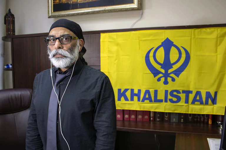 A Sikh man with a white beard stands in front of a yellow flag featuring a khanda above the word Khalistan