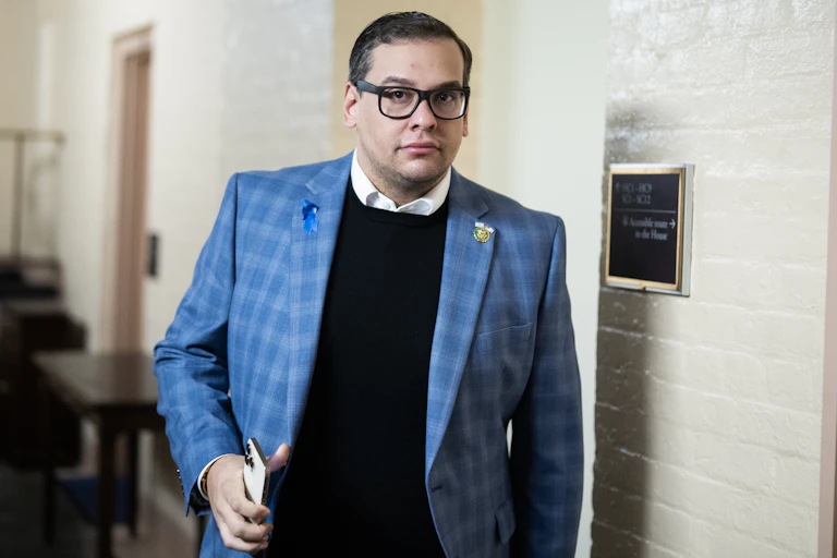 A dark haired man wearing glasses and a blue checked blazer over a dark sweater and white shirt.
