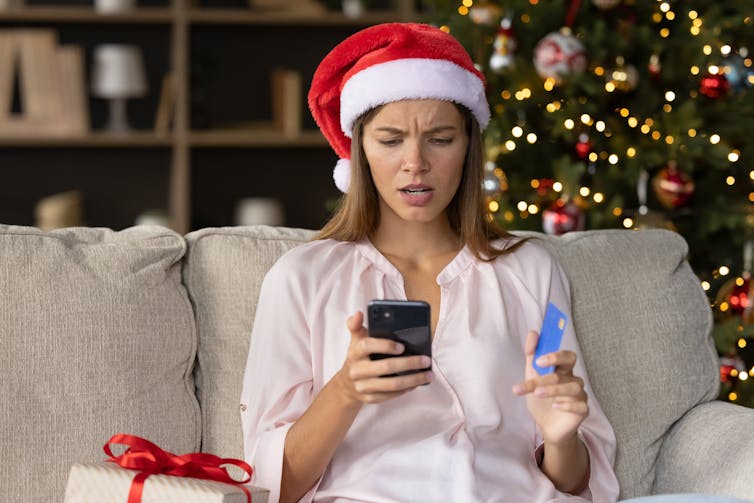 A woman in Santa hat looks shocked looking at her cellphone while holding a card.
