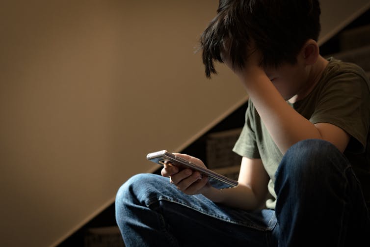 A boy sitting on stairs with his head in his hand, looking at a phone