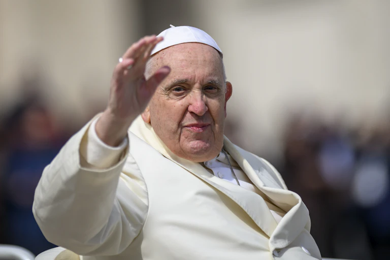 Pope Francis, dressed in white priestly garments, raises his hand to wave to people.