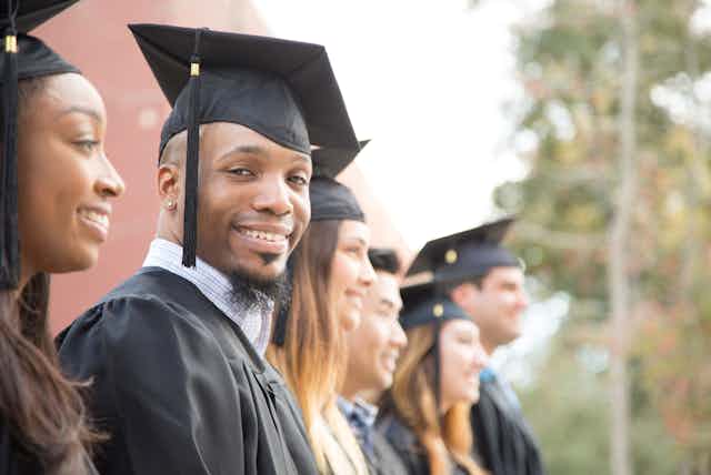 A Black man in a black graduation cap and gown smiles alongside other graduates.