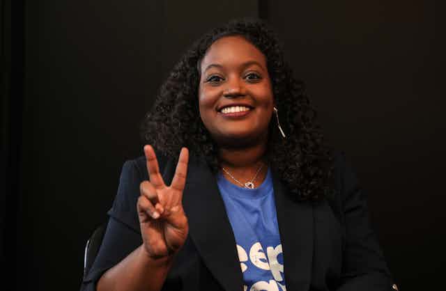 A Black woman in a dark suit jacket makes a victory sign with her right hand