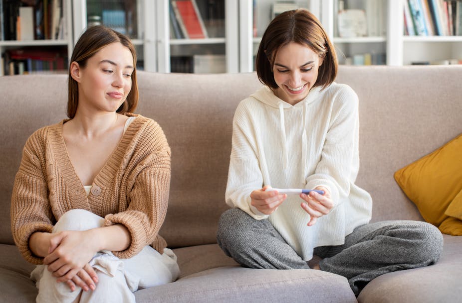 Two women sit on a sofa, one is smiling at a pregnancy test while the other looks disapprovingly at her