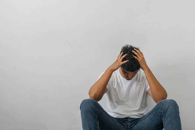 A dejected young man wearing a white T-shirt and blue jeans sits against a white wall with his hands on his head.
