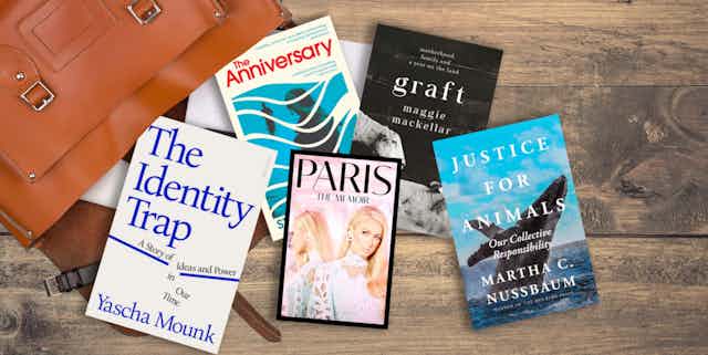 Five books spilling out of a brown leather bag onto a table. The books are The Identity Trap, Paris A Memoir, The Anniversary, Graft, and Justice For Animals.