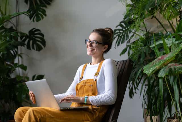 A young woman wearing yellow overalls sits with a laptop on her lap and several large plants behind her. She is laughing and looking at the computer.