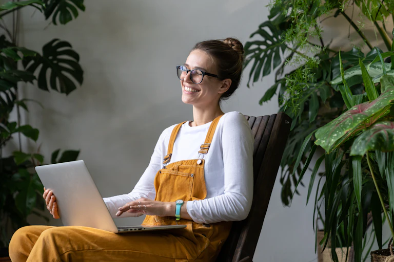 A young woman wearing yellow overalls sits with a laptop on her lap and several large plants behind her. She is laughing and looking at the computer.