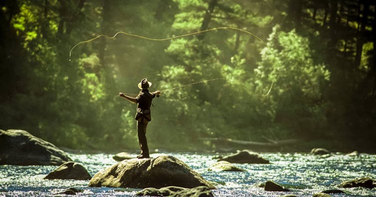 A man stands on rocks and casts a fly-fishing rod in a body of water surrounded by trees.