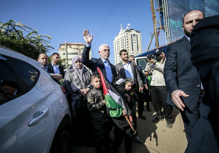 A man with grey hair salutes a crowd of people as he gets out of a car.