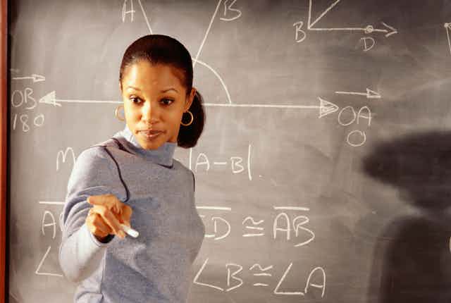 A female math instructor is at a chalkboard that has some algebraic equations written on it. She is pointing at someone who is not seen.