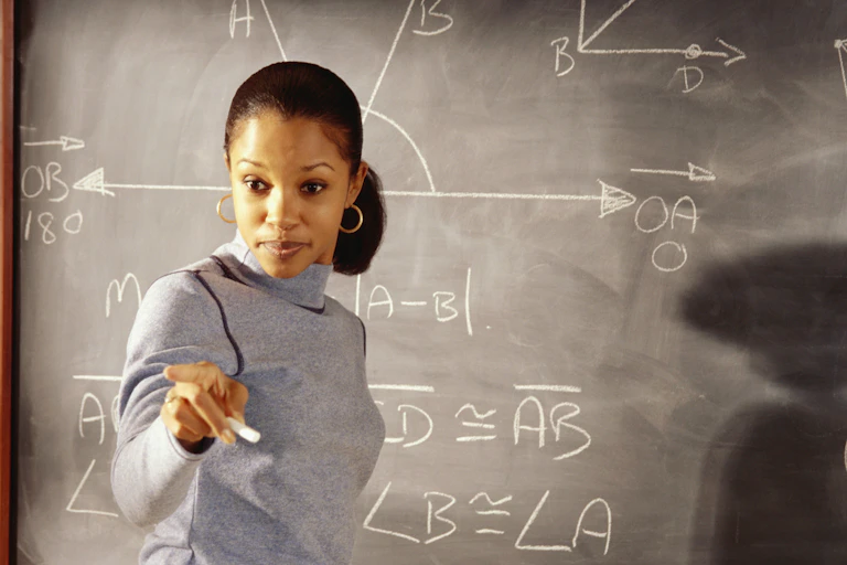 A female math instructor is at a chalkboard that has some algebraic equations written on it. She is pointing at someone who is not seen.