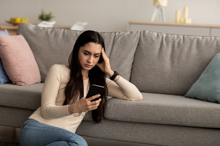 Woman leans on sofa while looking at phone with stressed expression.