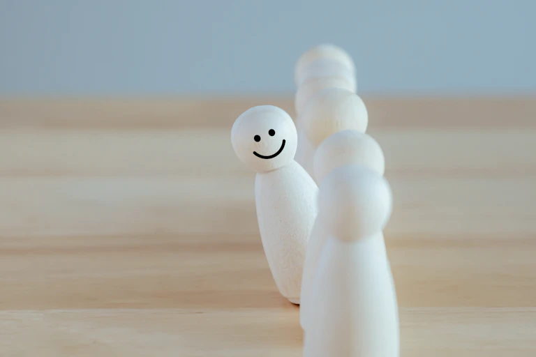 A line of wood pegs. One is peeking out from the others with a smile drawn on the head of it.