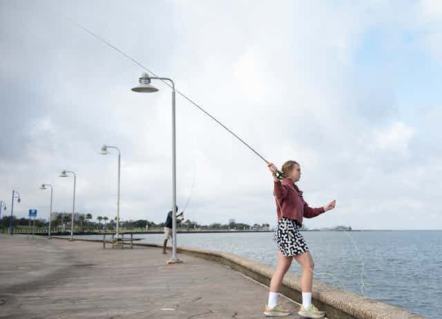 A female student holds a fly-fishing rod while standing on a walkway near a lake.