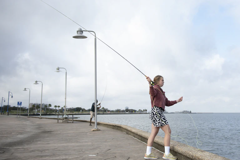 A female student holds a fly-fishing rod while standing on a walkway near a lake.