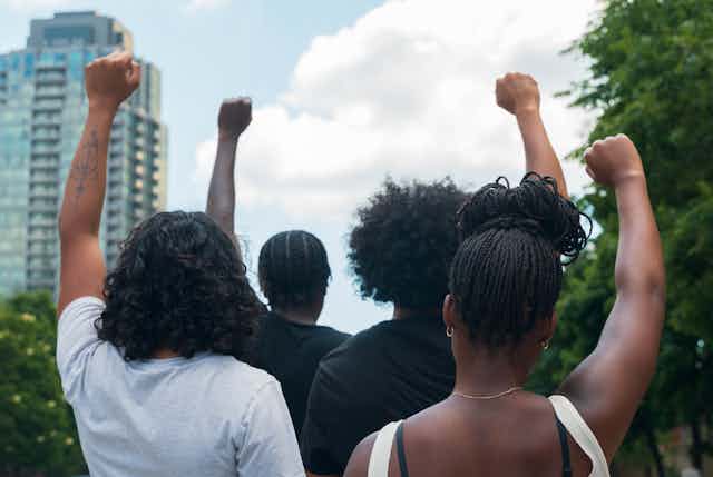 Two men and two women holding up their hands in a fist in protest, with their backs to the camera and a highrise building in front of them.