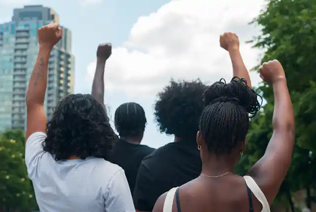 Two men and two women holding up their hands in a fist in protest, with their backs to the camera and a highrise building in front of them.