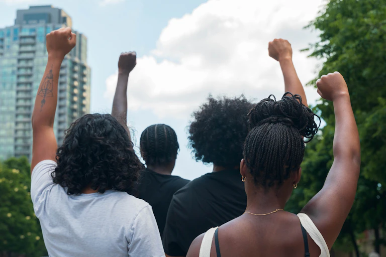 Two men and two women holding up their hands in a fist in protest, with their backs to the camera and a highrise building in front of them.