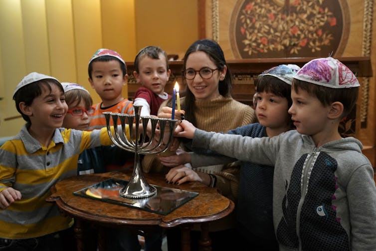 A brunette woman crouches indoors by a table, amid a handful of children, as she holds a lit candle.