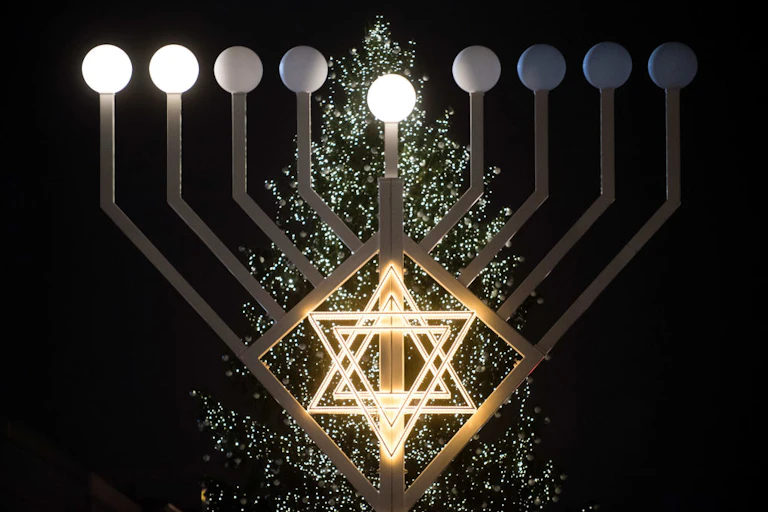 Lights in the shape of a six-pointed star positioned in front of large lamppost-like structures and a decorated tree.