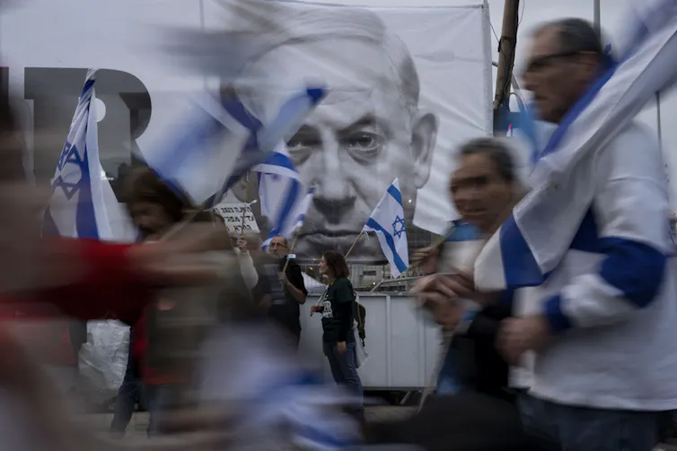 A poster of Benjamin Netanyahu behind a blurred crowd of people with Israeli flags.
