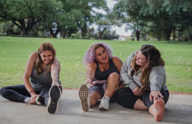 Three people are sitting on the ground wearing activewear and stretching while laughing.