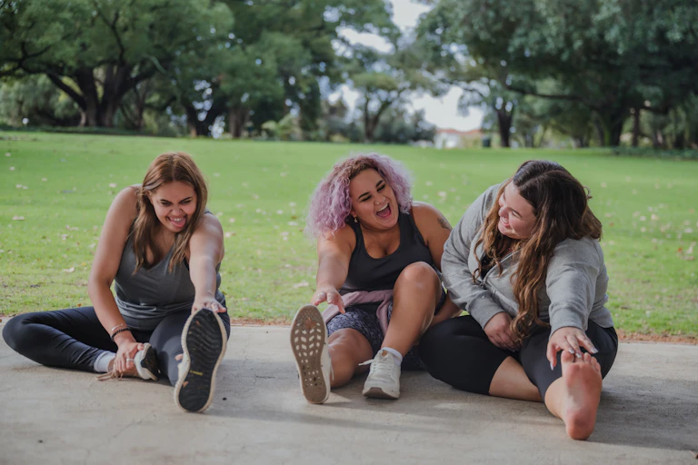 Three people are sitting on the ground wearing activewear and stretching while laughing.