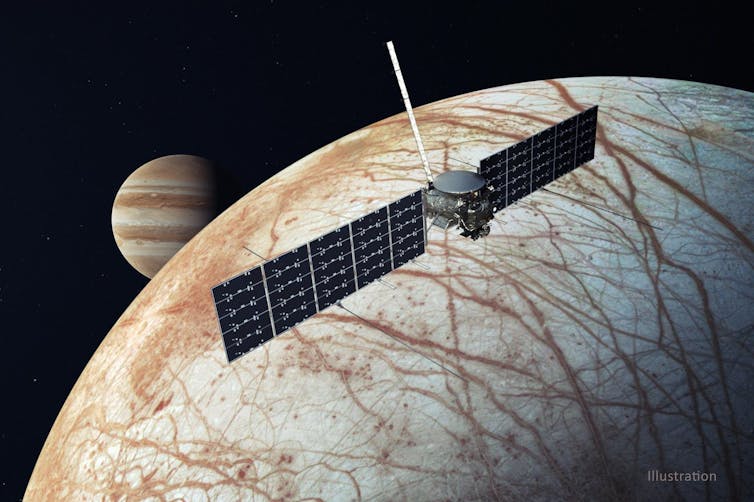 Una nave espacial con dos grandes paneles rectangulares que salen de un pequeño cilindro vuela por encima de una luna marrón y blanca, con un planeta a rayas marrones al fondo.