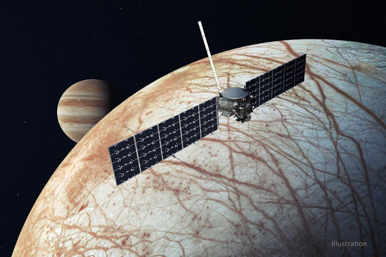 Uma nave espacial com dois grandes painéis retangulares emergindo de um pequeno cilindro voa sobre uma lua marrom e branca, com um planeta com listras marrons ao fundo.