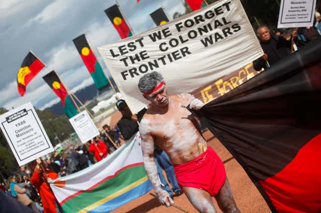 A sunny day in Canberra, people walk with flags, including many Aboriginal flags. There is a person wearing a red headband and lap-lap painted up in white ochre who carries a large Aboriginal flag like a banner. Behind them is a sign that reads 'Lest we forget the colonial frontier wars'.