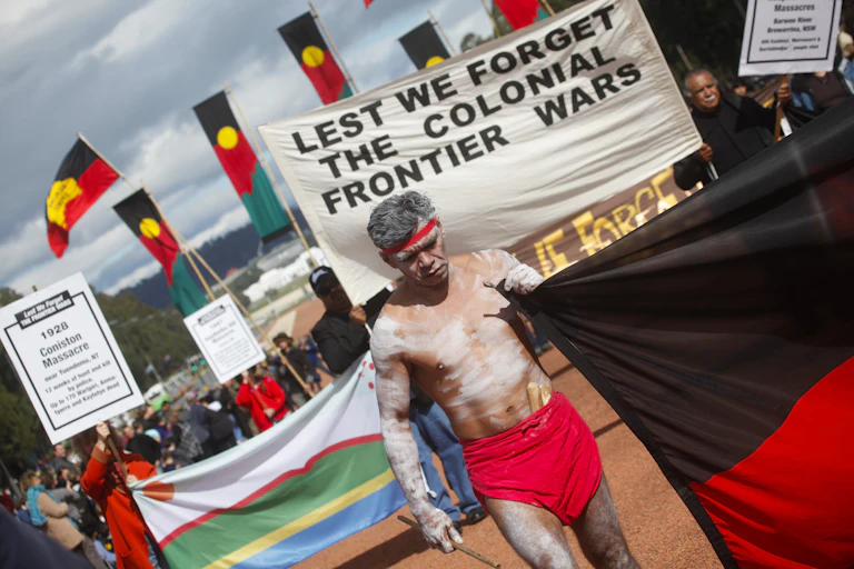 A sunny day in Canberra, people walk with flags, including many Aboriginal flags. There is a person wearing a red headband and lap-lap painted up in white ochre who carries a large Aboriginal flag like a banner. Behind them is a sign that reads 'Lest we forget the colonial frontier wars'.