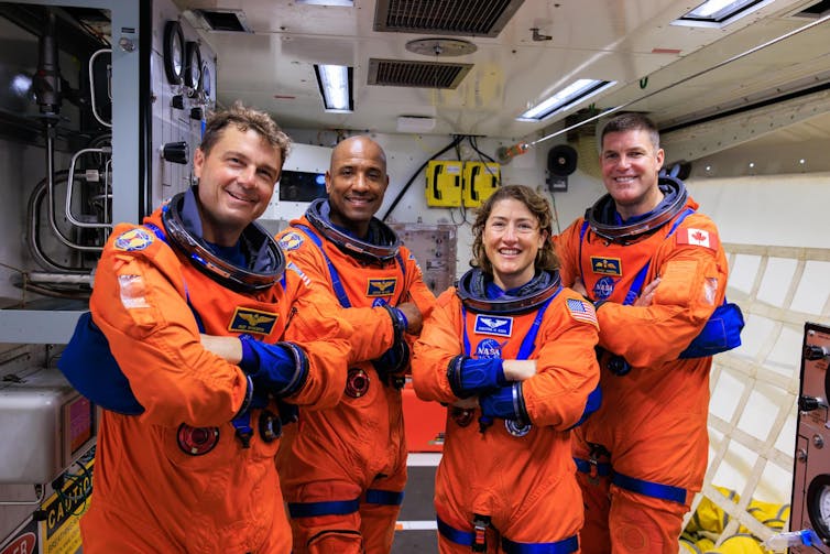 Quatro pessoas em trajes espaciais laranja em uma pequena sala branca.