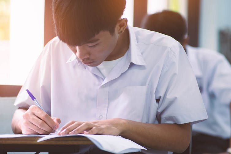 A boy in school uniform writes with a blue pen into an exercise book.