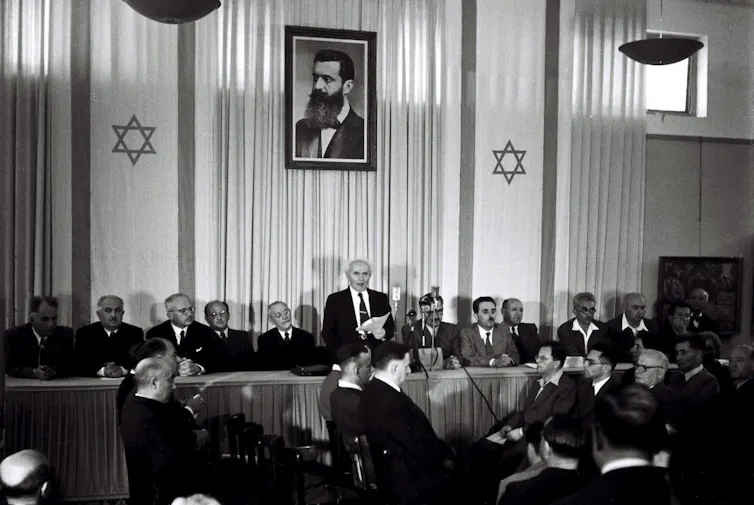 Black and white photo of a row of men sitting behind a long table facing a crowd of men