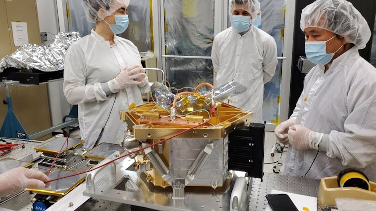 Three scientists wearing lab coats standing around a table which has a metal instrument half-assembled on it.