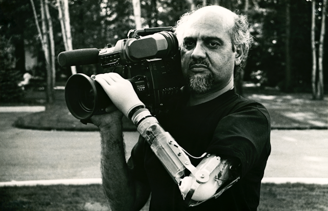 A balding man holds a camera on his shoulder with two arms - one of them is a prosthetic, bionic arm with mechanics and wires exposed.