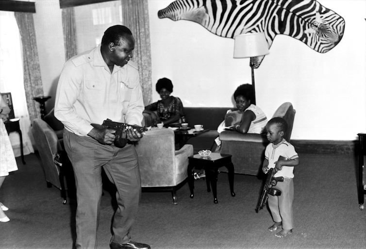 A man holds a gun as he plays with a little boy, also holding a gun, in a lounge. In the background, women are seated and a zebra skin adorns the wall.