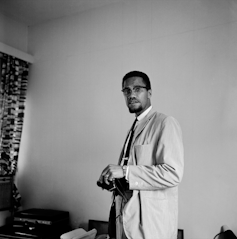 A man in glasses and a jacket stands looking at the camera in a hotel room.