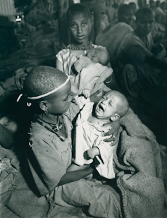 Un niño sostiene a un bebé que llora mientras una niña demacrada mira directamente a cámara. Detrás de ellos hay gente sentada en el suelo.