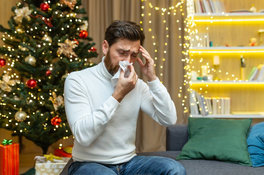 A man blows his nose into a tissue. A Christmas tree is in the background behind him.