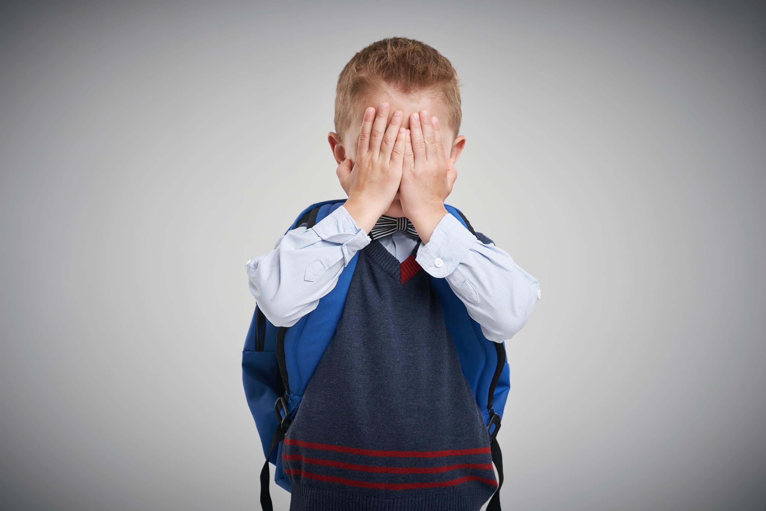 A photograph of a boy dressed for school, wearing a backpack, and covering his face
