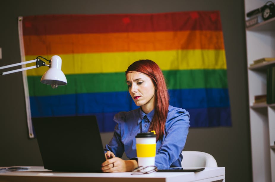 A young office worker sits at a desk with a laptop and coffee. A pride flag is hung on the wall.