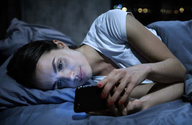  Young tired female looking at her mobile phone screen, lying in bed late at night,