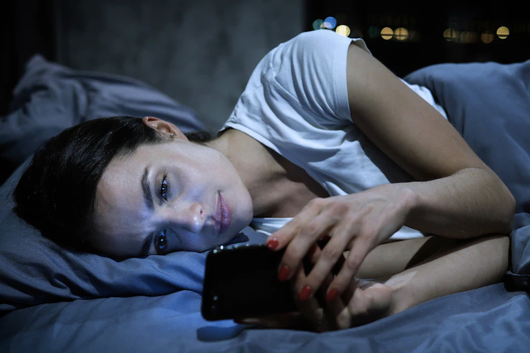 Young tired female looking at her mobile phone screen, lying in bed late at night,