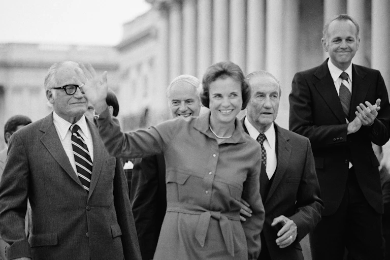 A smiling woman in a shirtwaist dress waves to the public while standing in front of four men.
