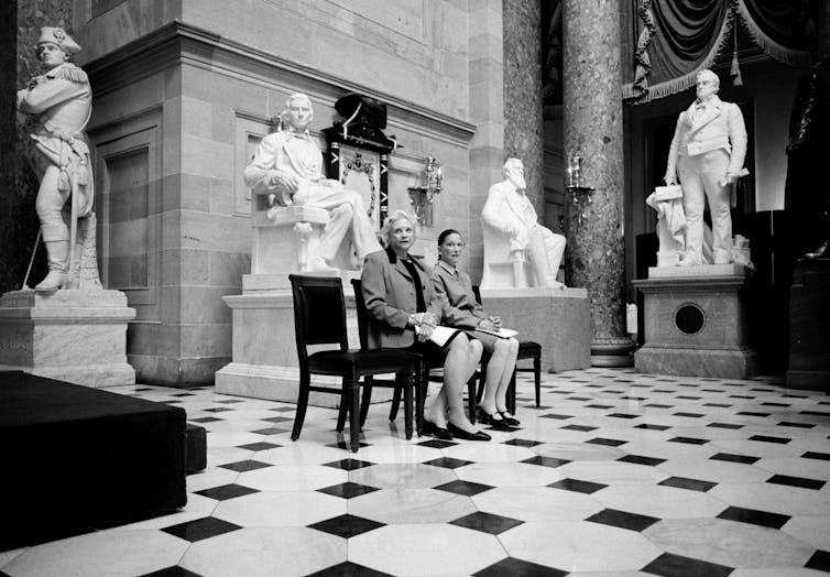 Dos mujeres sentadas en un gran salón con suelo de mármol, rodeadas de grandes estatuas de hombres.